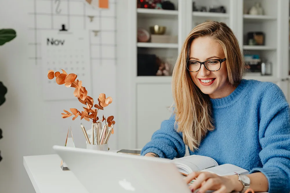 A smiling woman with glasses working on a laptop and notebook in a home office, illustrating the process of planning and managing FSA or HSA contributions for health expenses.
